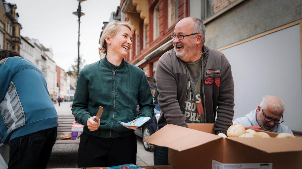 Ines Schwerdtner und Mathias Baum stehen zur Küche für alle hinter eine, Tisch und lachen.