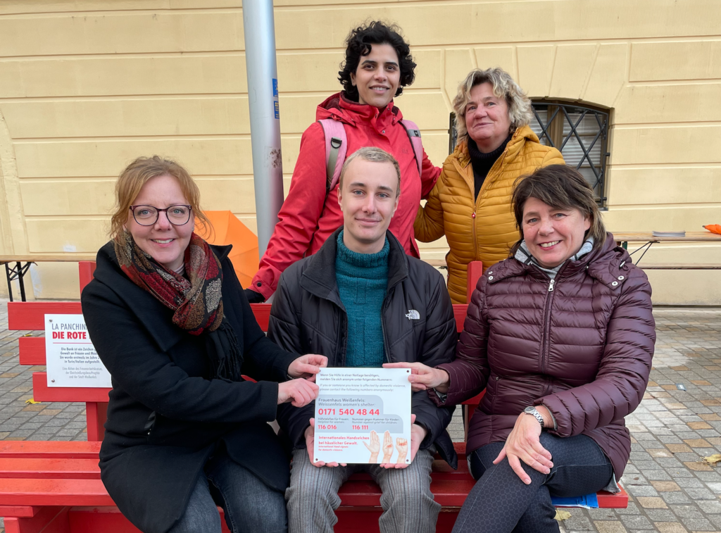 Zu sehen sind fünf Personen. Zwei Frauen und ein Mann sitzen im Vordergrund auf der Roten Bank, die auf Gewalt an Frauen aufmerksam machen soll. Die Personen auf der Bank halten eine Plakette mit Informationen zur Bank in der Hand. Hinter der Bank stehen zwei weitere Frauen.